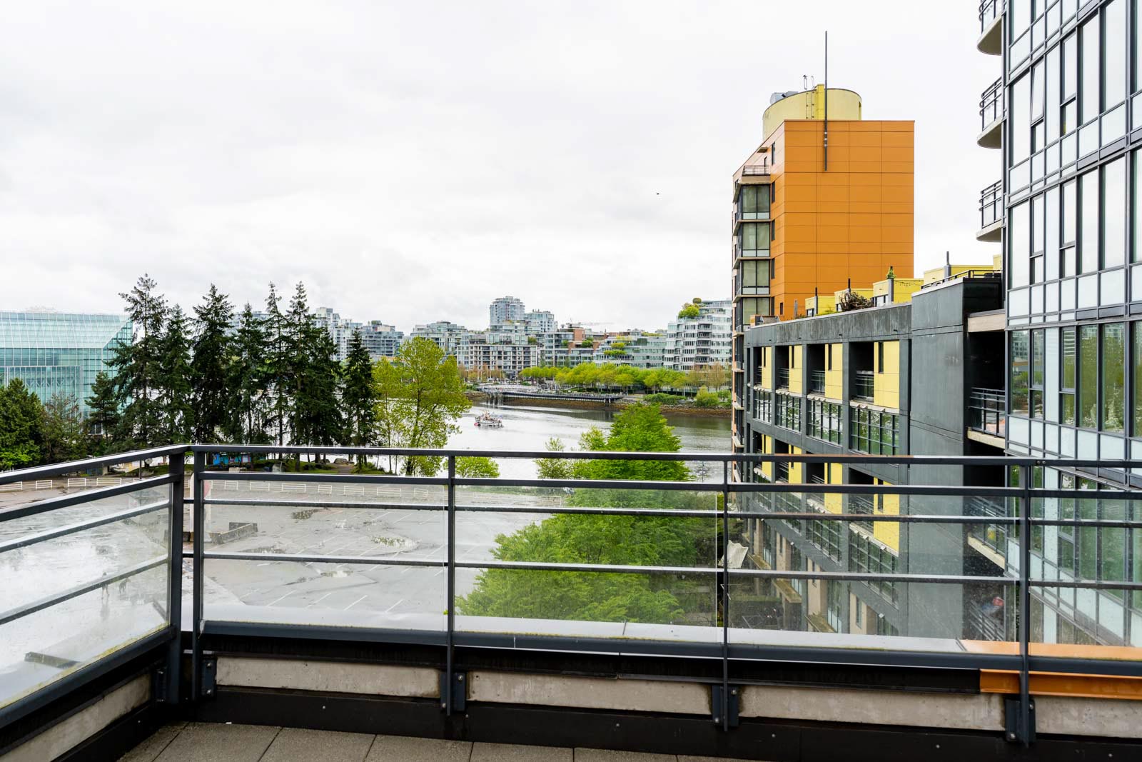 View from a balcony overlooking a river and cityscape with modern buildings, trees, and a cloudy sky.