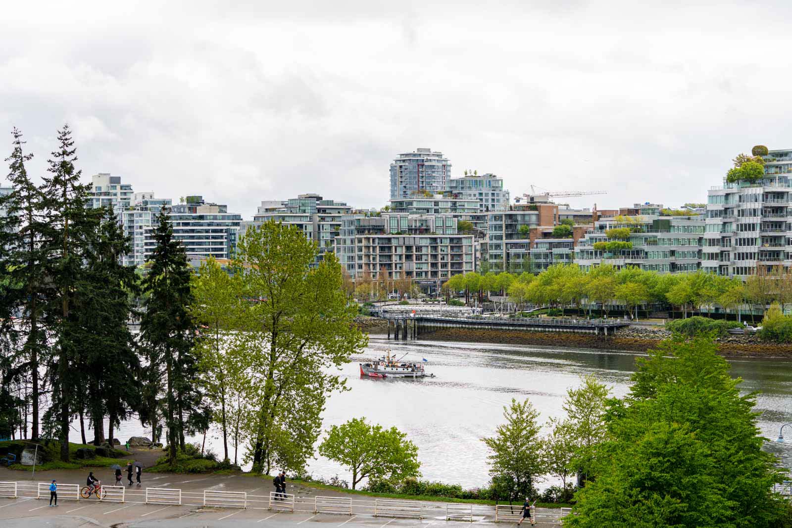 A river with a small boat floats through a cityscape lined with modern buildings and trees under a cloudy sky.