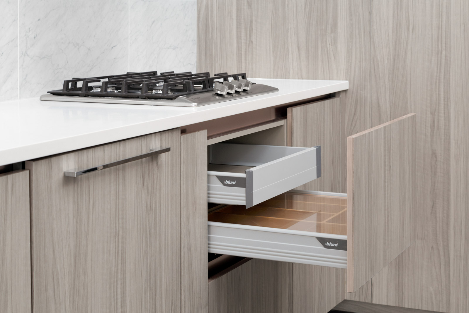 A modern kitchen with a gas cooktop on a white countertop and two open drawers in a light wood cabinet below.