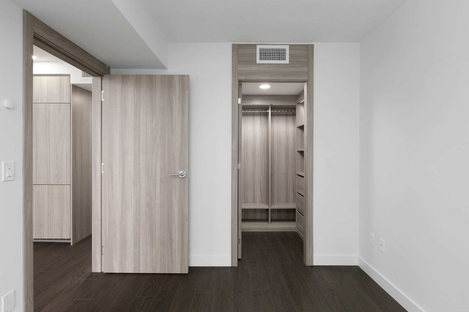 A modern, empty room with light wood doors, white walls, dark wood flooring, and an open walk-in closet with shelves and a bench.
