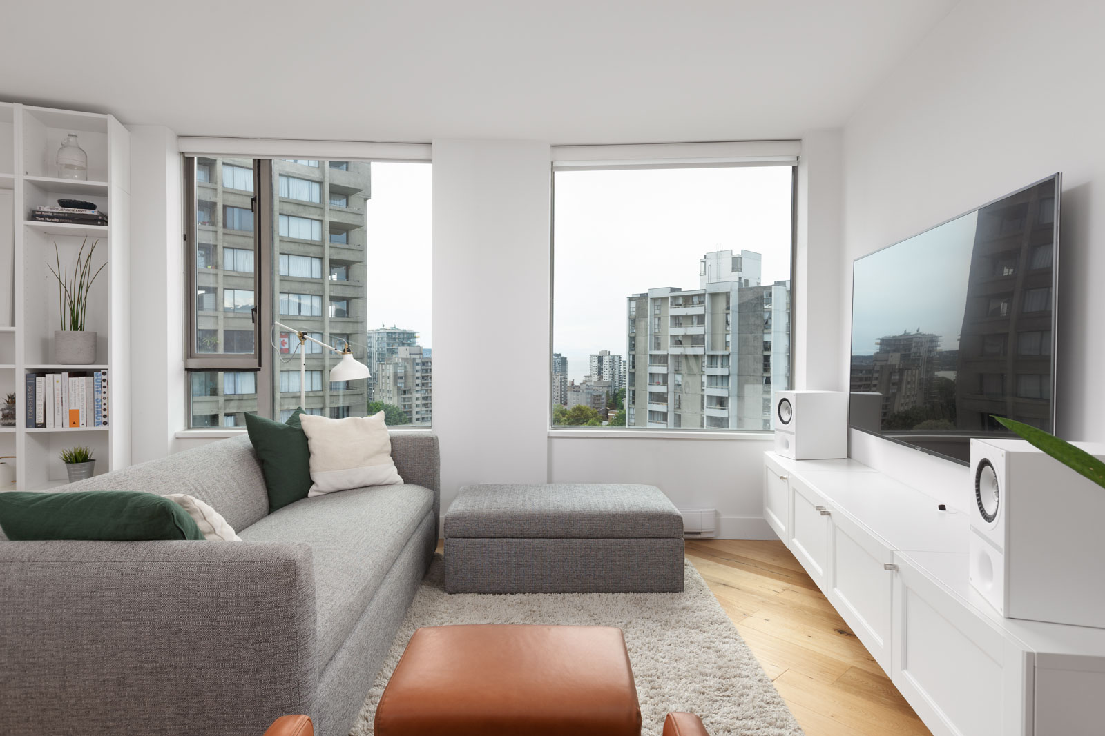 Modern living room with a grey sofa, ottoman, white TV console, large flat-screen TV, city view through tall windows, and shelves with books and decor.