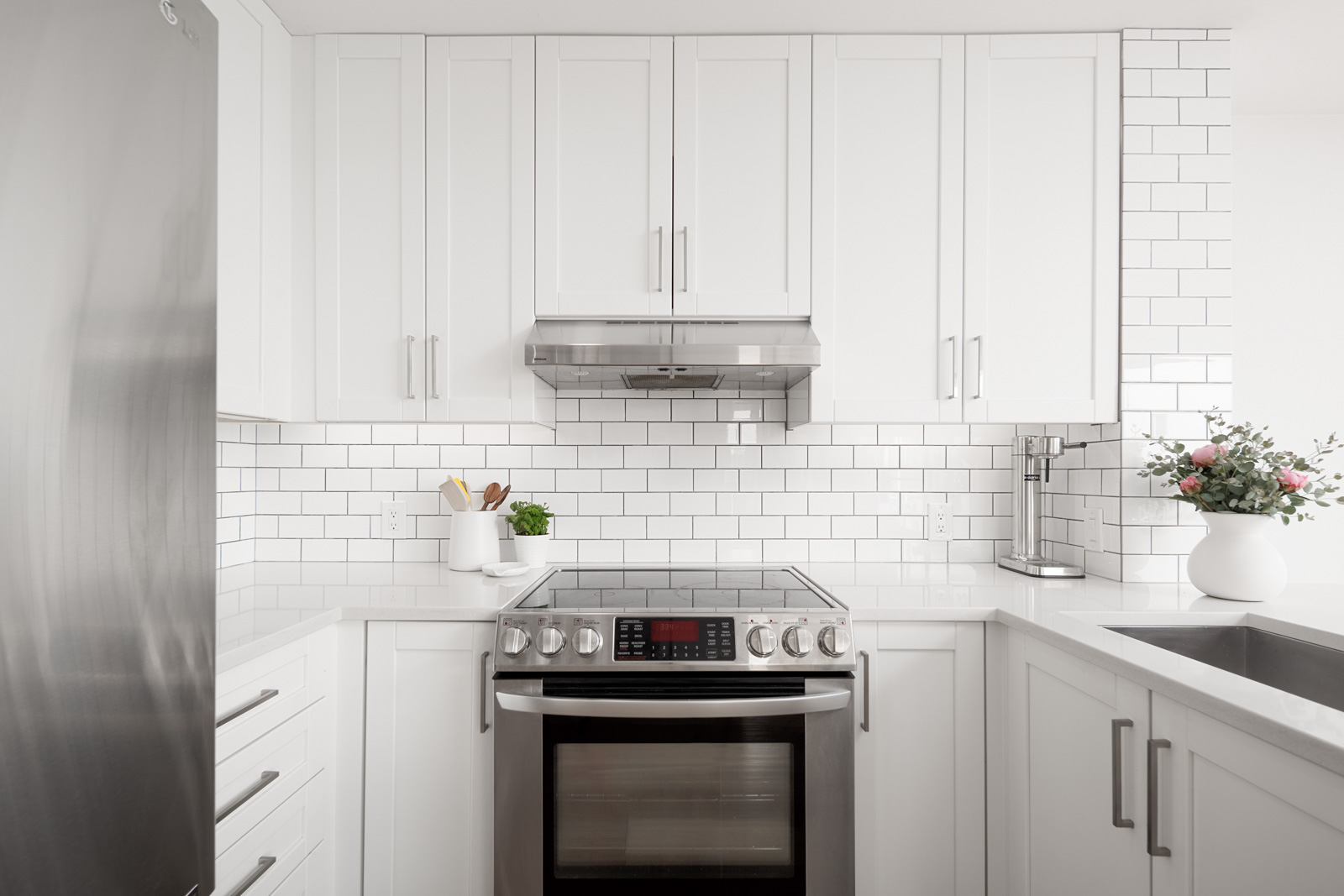 Modern white kitchen with stainless steel appliances, white subway tile backsplash, potted plant, utensils, and a vase of flowers on the counter.