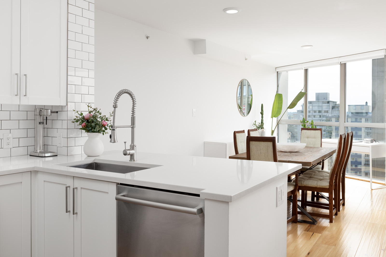 Modern kitchen with white cabinets, stainless steel appliances, a vase with flowers on the counter, and a dining area with large windows in the background.