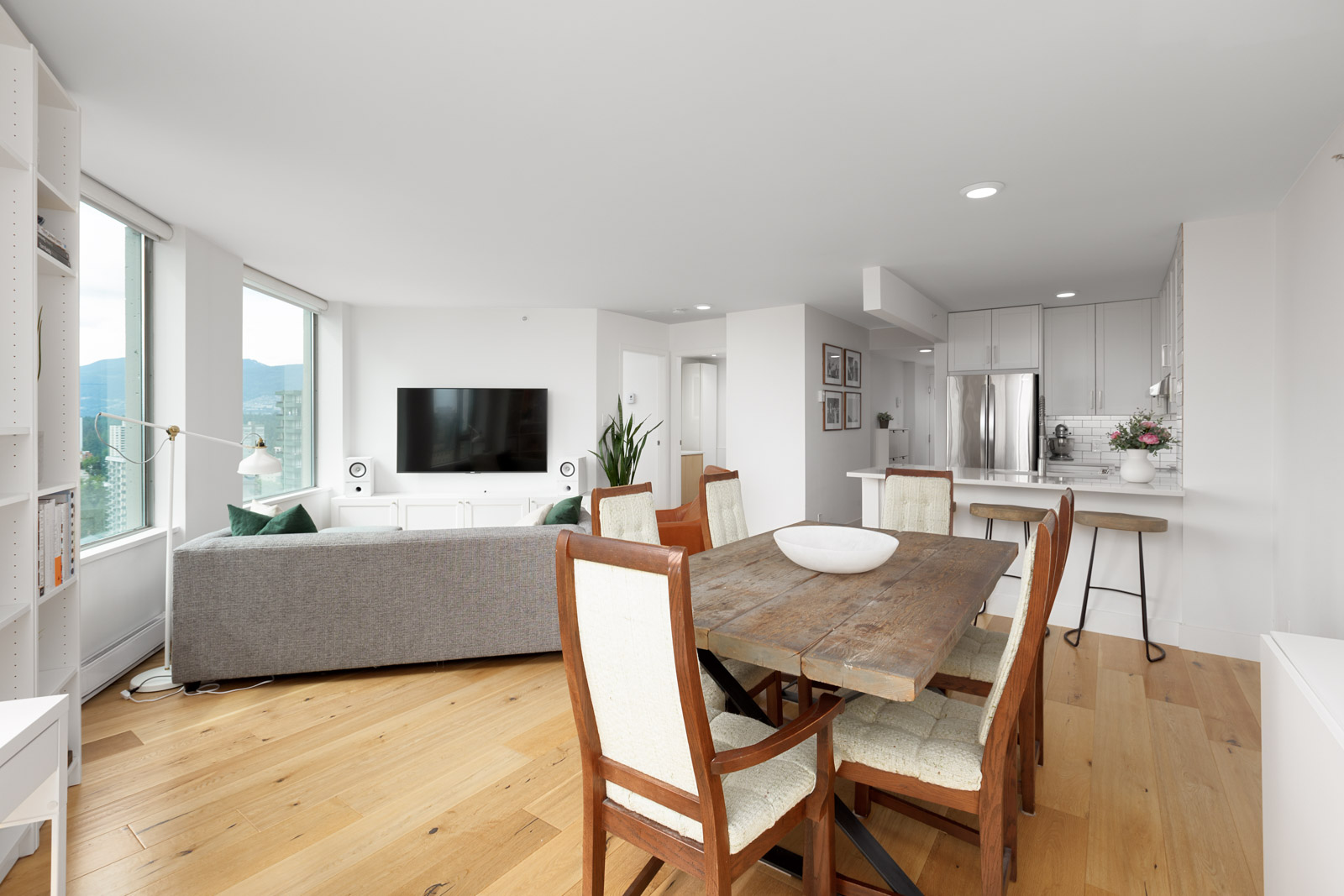 Open-concept living and dining area with light wood flooring, a gray sofa, wall-mounted TV, rustic dining table, and modern white kitchen with stainless steel appliances.