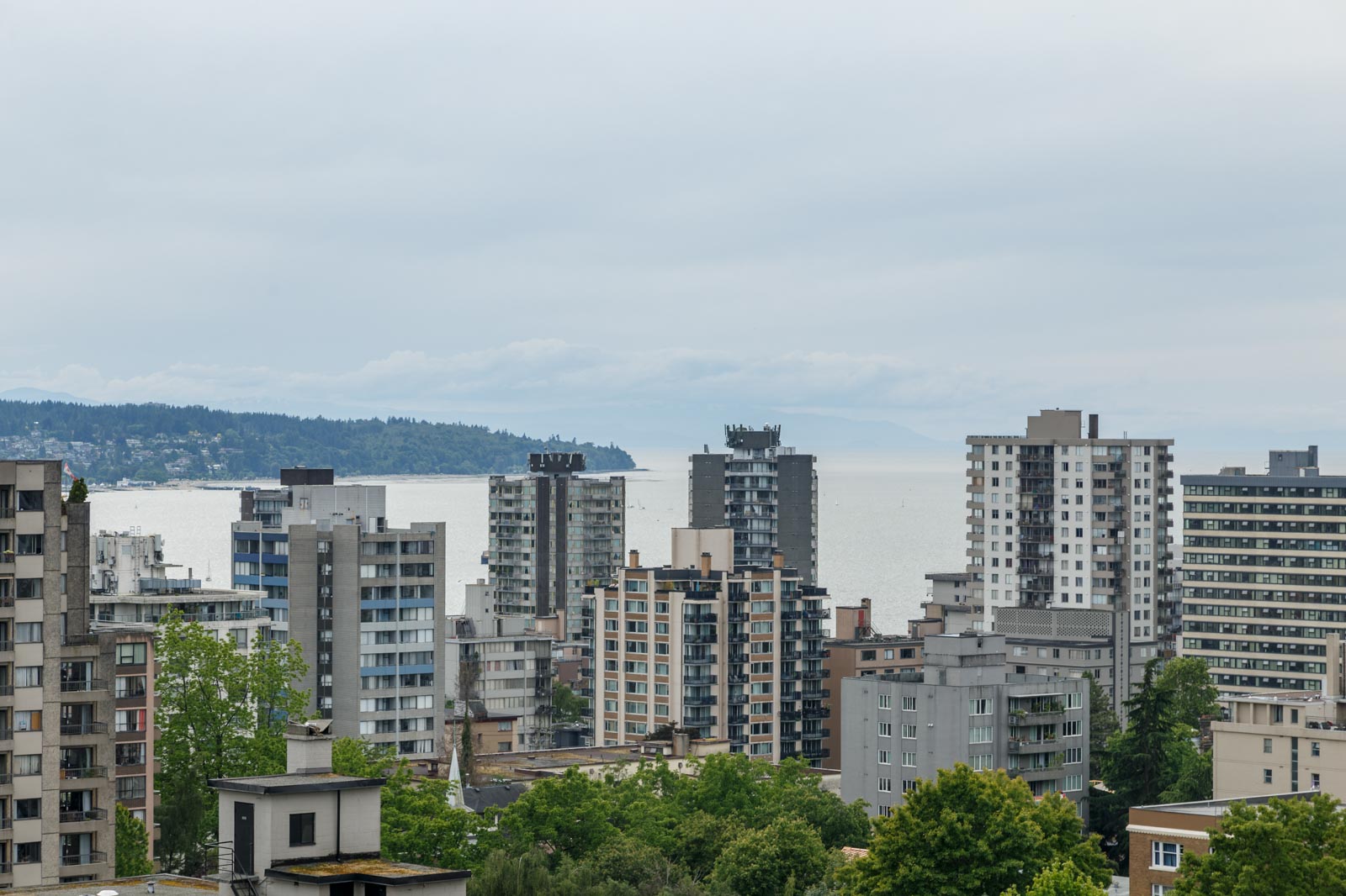 High-rise apartment buildings with a view of the ocean in the background under a cloudy sky, surrounded by greenery and distant hills.