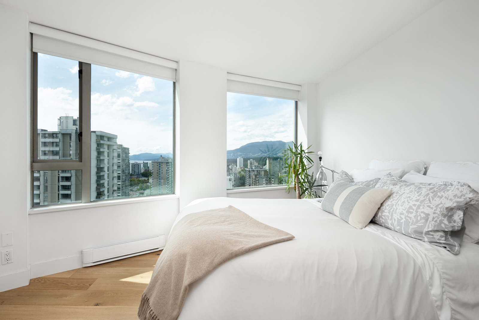 A modern, white bedroom with large windows offering city and mountain views, featuring a neatly made bed with decorative pillows and a beige throw.