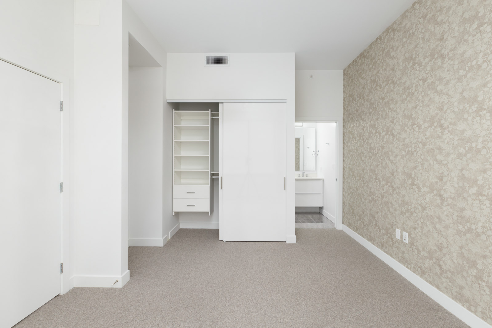 Empty room with beige carpet, open closet with shelves and drawers, and a view into a white bathroom. One wall is covered with patterned wallpaper.