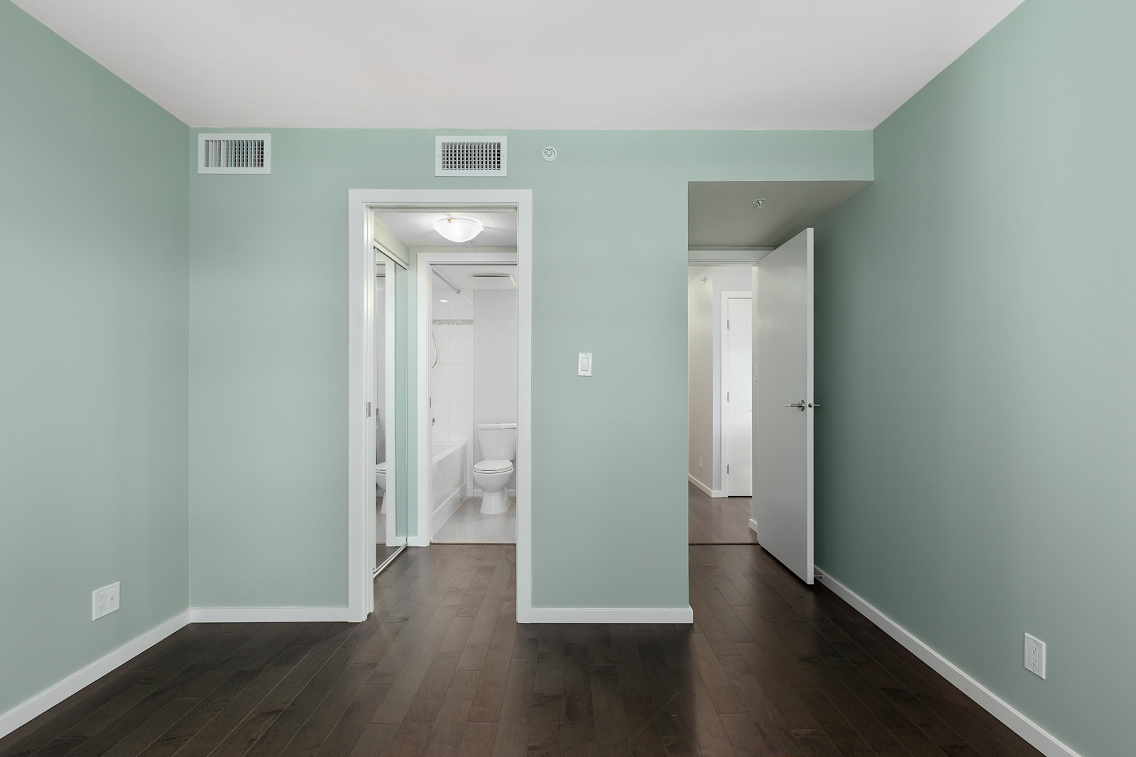 Empty bedroom with light green walls, dark wood floor, open doorways leading to a bathroom with white fixtures, and another to a hallway.