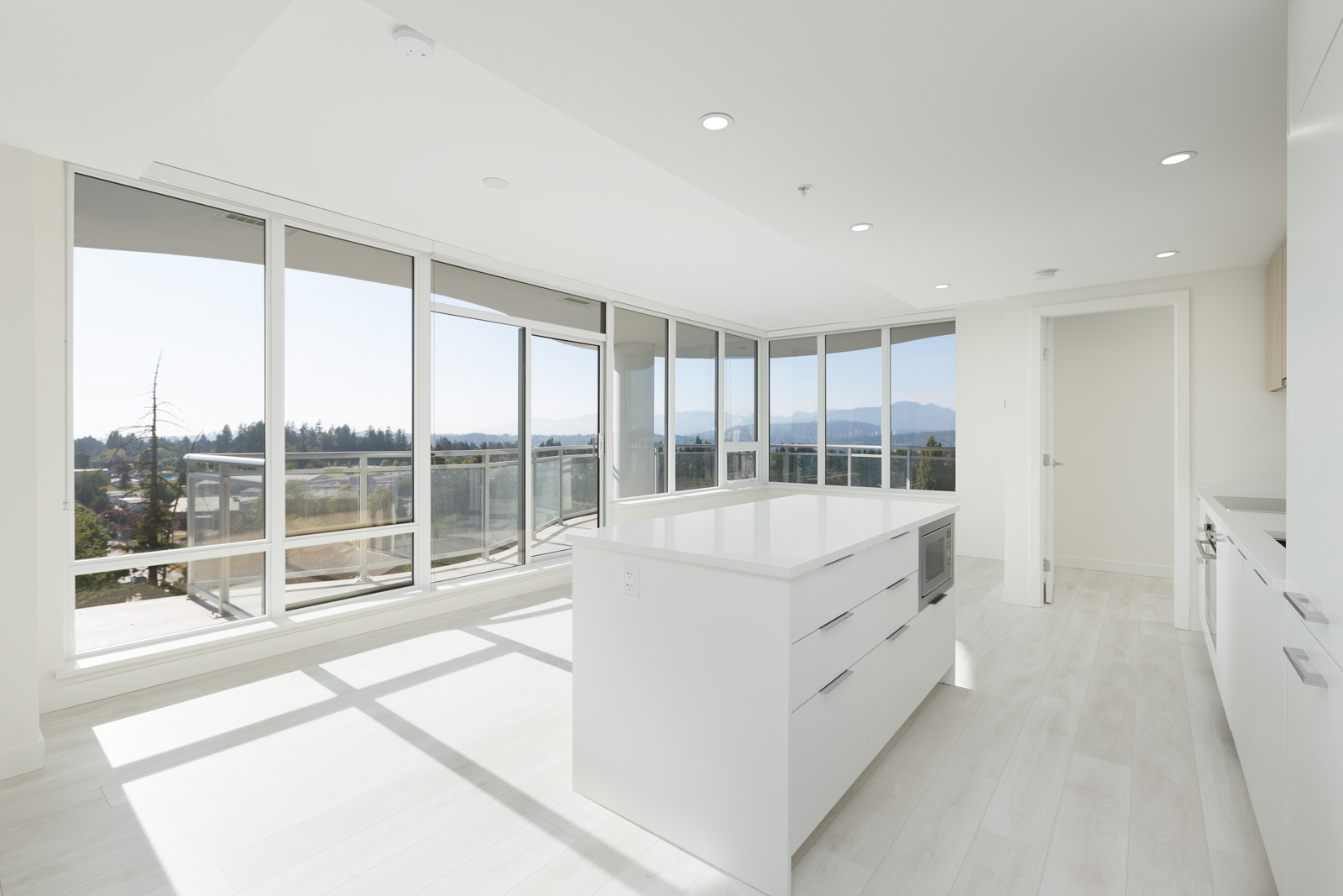 Bright modern kitchen with white cabinetry, central island, and large floor-to-ceiling windows overlooking a balcony and scenic view.