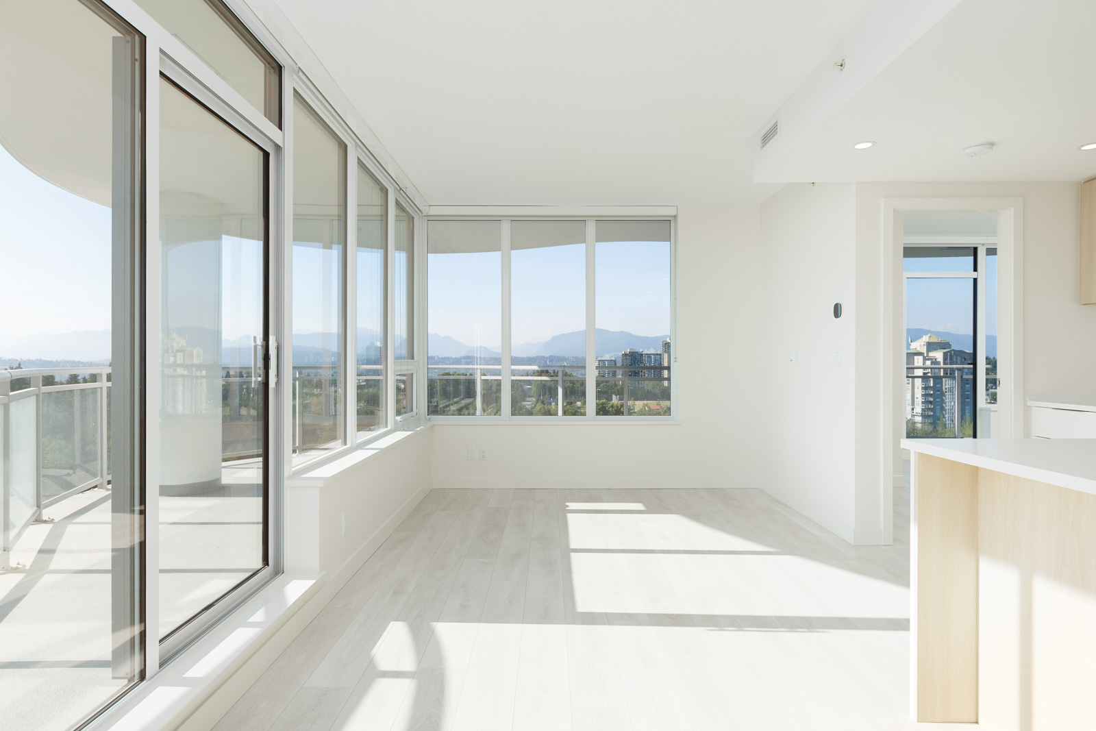 Bright, empty apartment with large windows and a balcony. The room features light wood flooring and white walls, offering a scenic view of the mountains and cityscape.