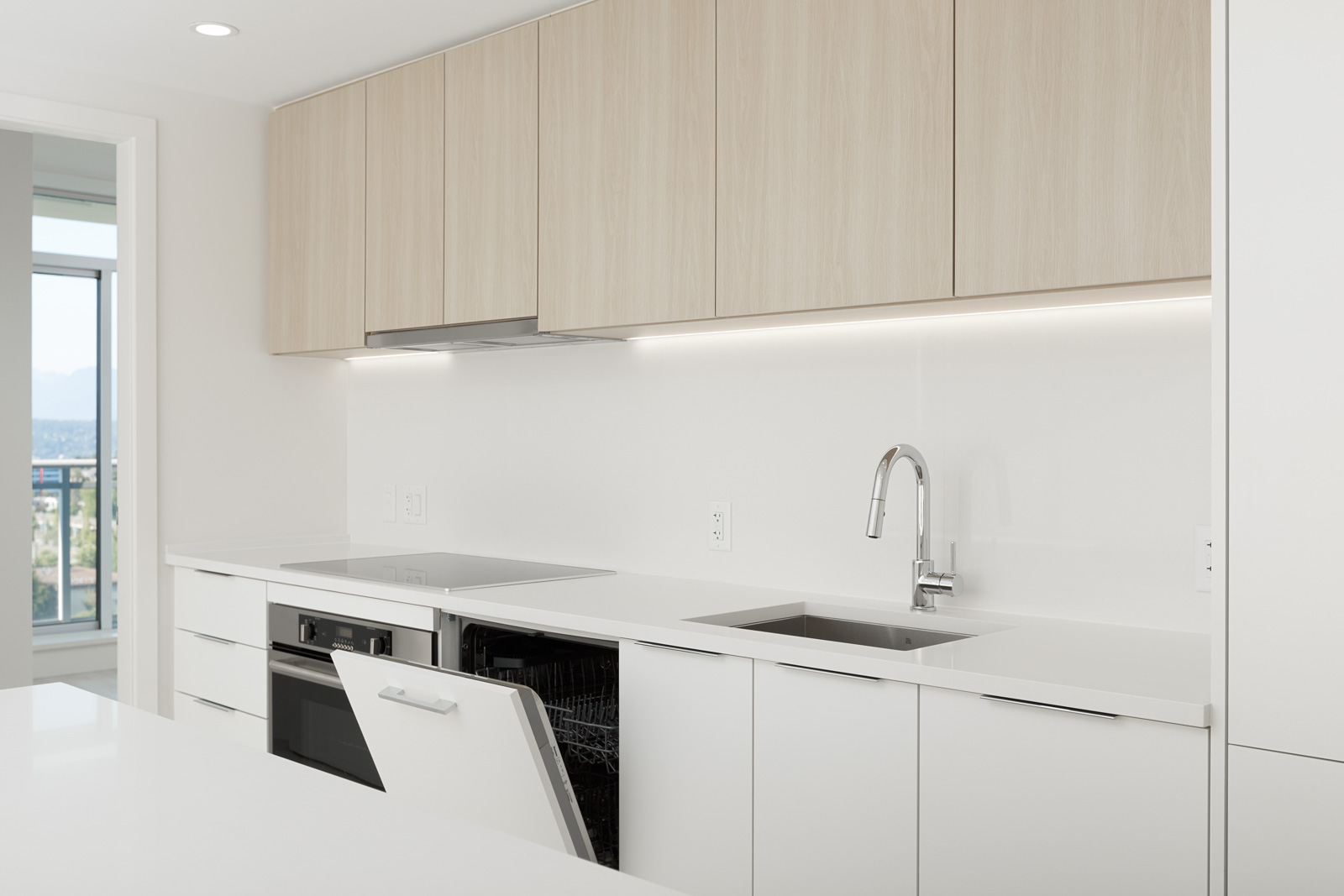 Modern kitchen with light wood cabinets, white countertops, and a stainless steel sink. An open dishwasher is visible under the counter. Large window in the background.