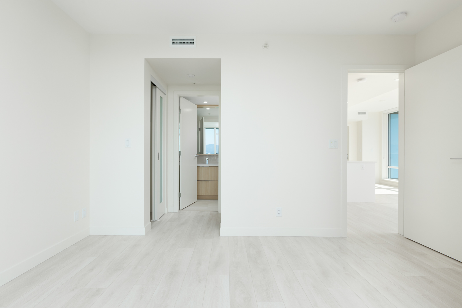 Minimalist empty white room with light wooden floors, an open doorway leading to a bathroom, and another open door showing a sunlit area.