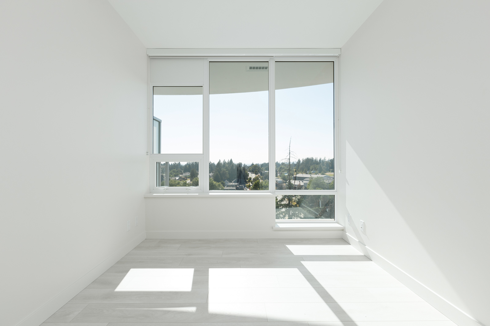 Bright, empty room with large windows offering a view of trees and sky. White walls and light wood flooring enhance the airy feel.