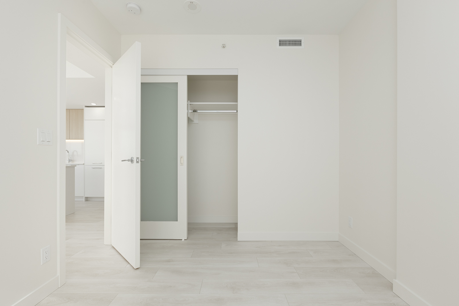 Empty room with a white door ajar, revealing a closet with sliding door. The room has white walls and light wooden flooring. A glimpse of a kitchen is visible through the doorway.