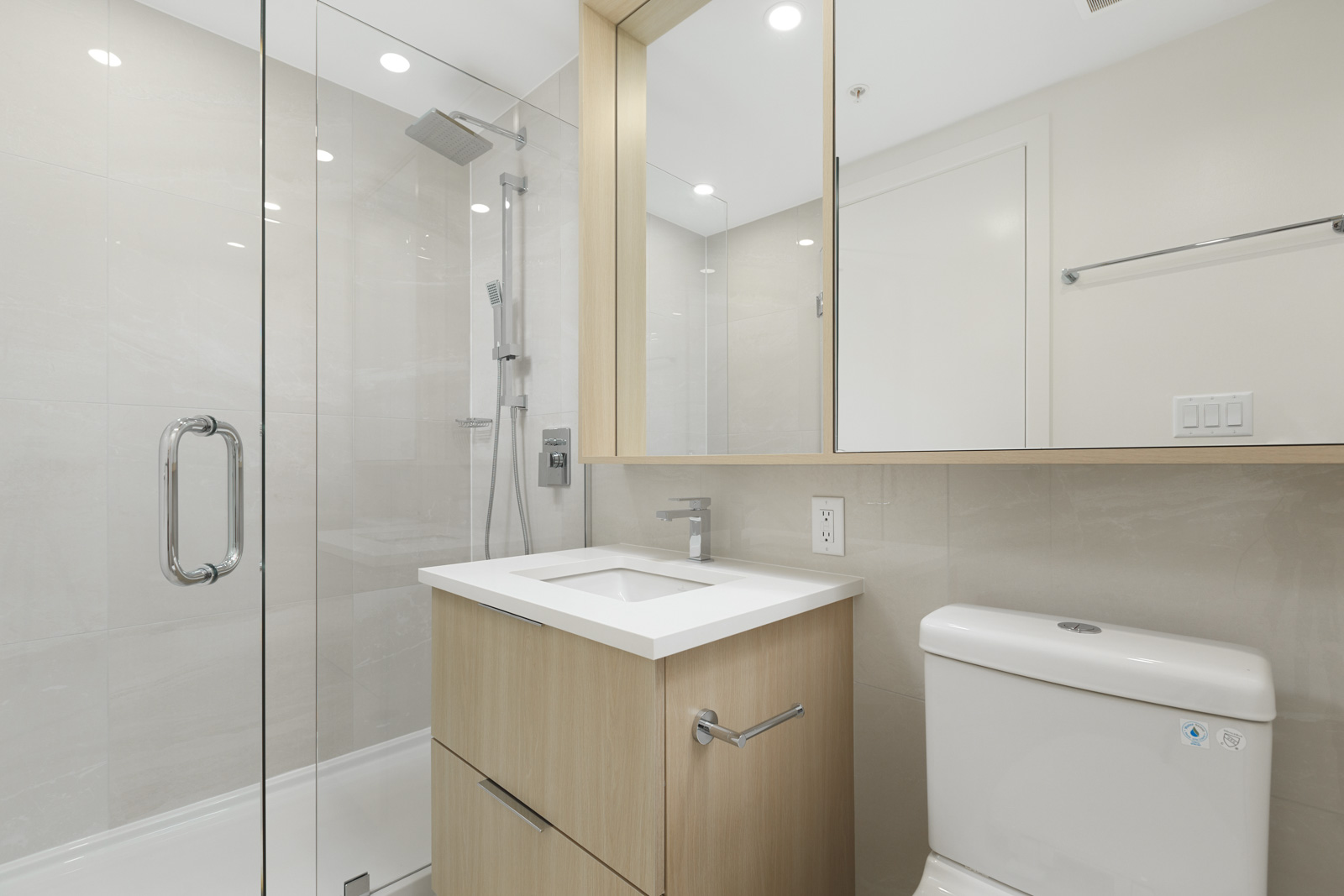 Modern bathroom with glass-enclosed shower, wooden vanity with sink, and white toilet. Beige tiled walls and a mirror cabinet above the sink.