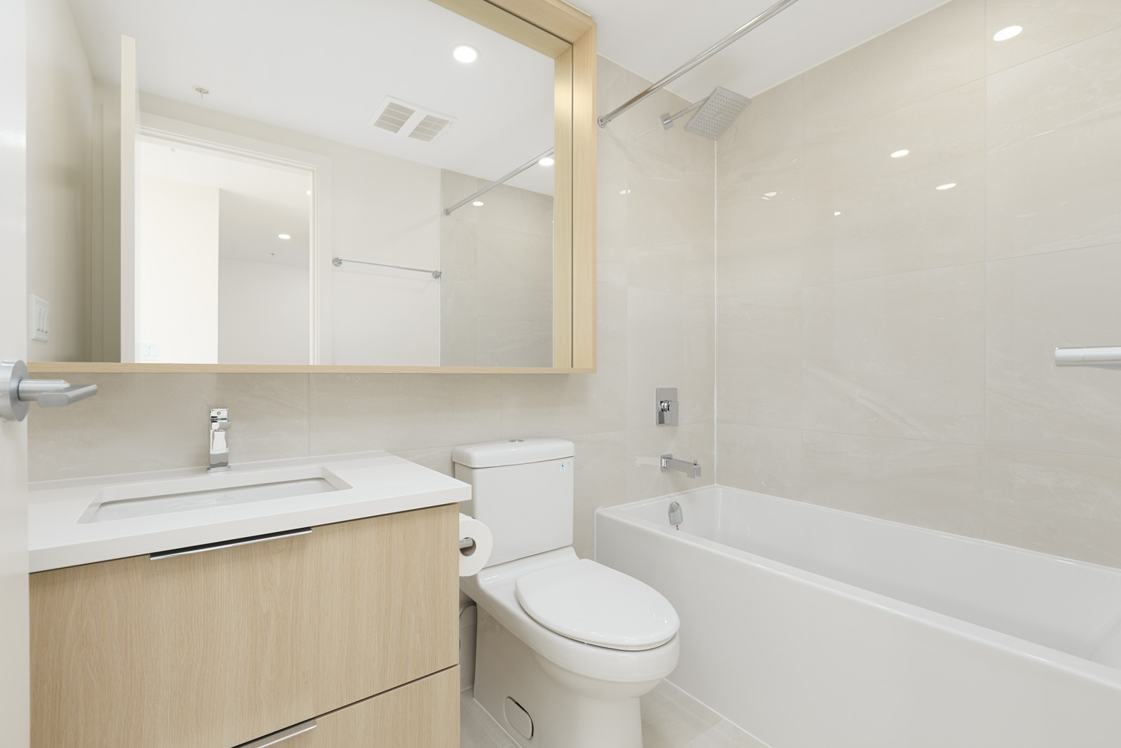 Modern bathroom with light wood cabinets, a white sink, toilet, and a bathtub with a showerhead. Large mirror above the sink. Walls and floor are tiled in light beige.