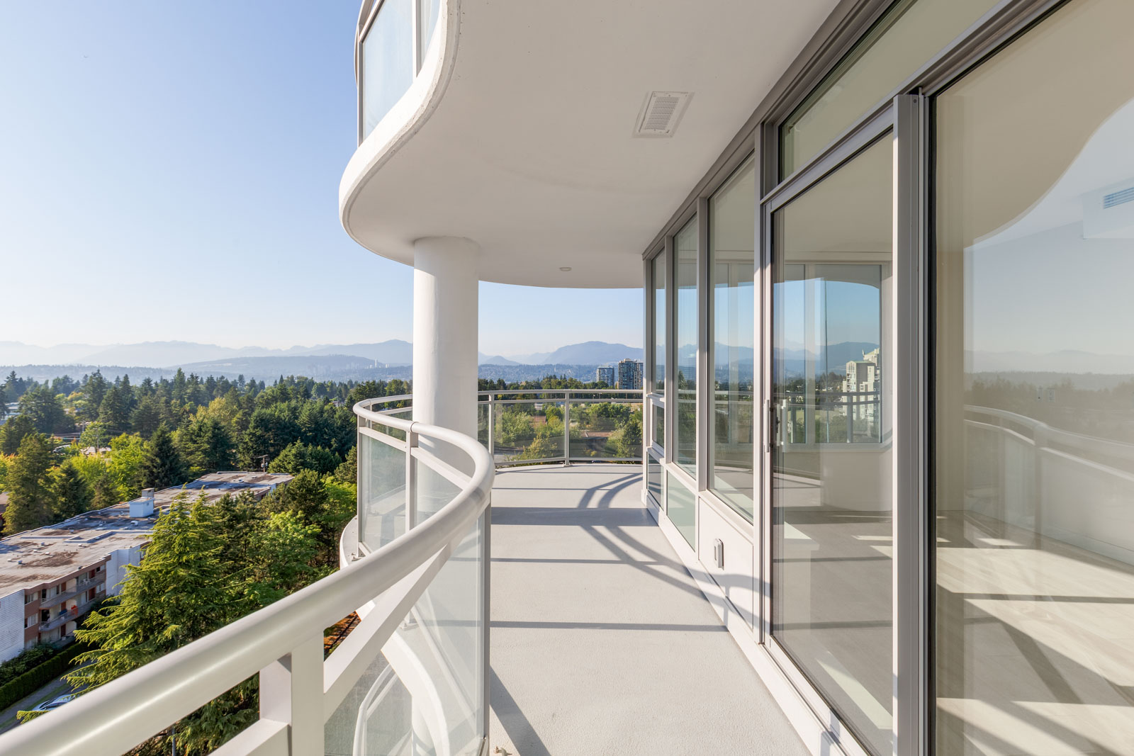 A modern balcony with glass railing offers a view of distant mountains, trees, and buildings under a clear blue sky.