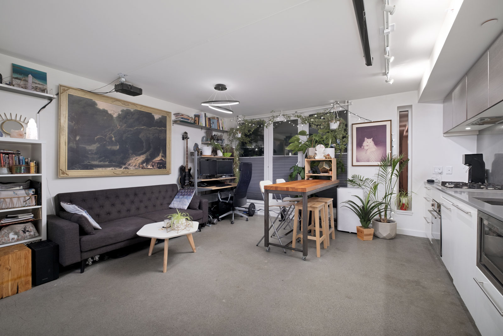 Modern living room and kitchen with a gray sofa, wall art, plants, small dining table, bookshelf, and a desk setup near a window.