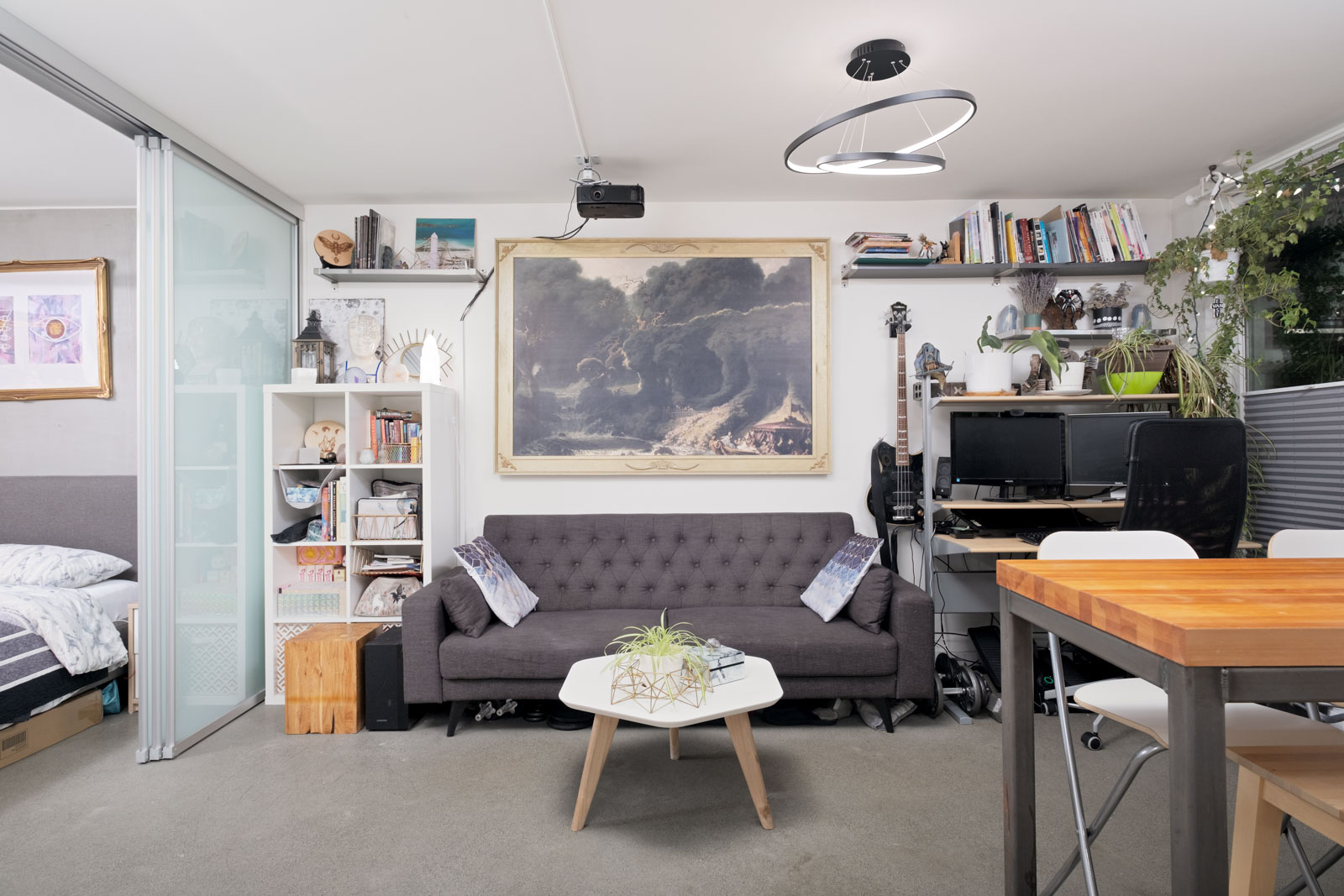 Modern studio apartment with a gray sofa, coffee table, art on the wall, bookshelves, a guitar, computer desk, and a dining table; bed area visible to the left.