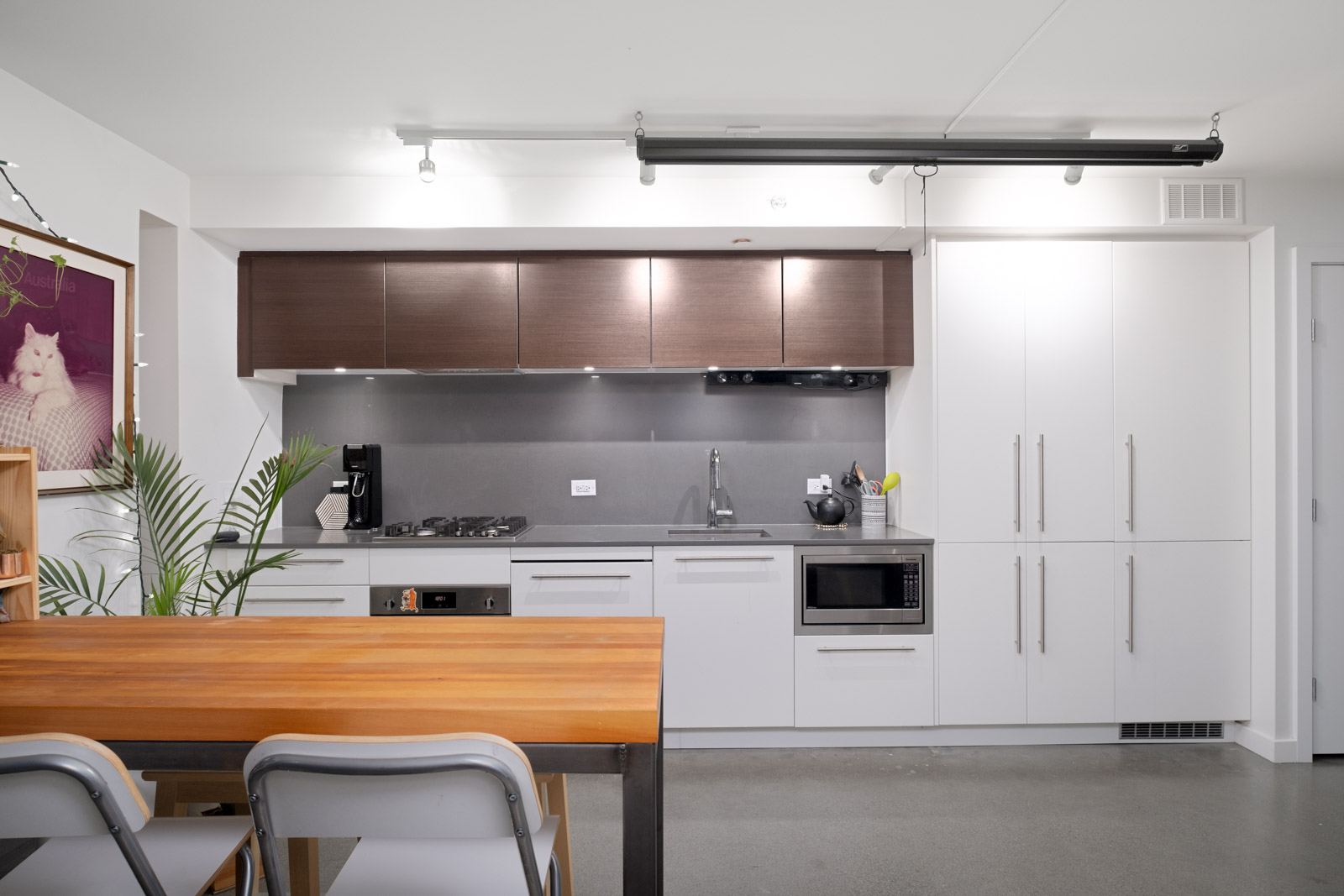 Modern kitchen with white cabinets, brown upper cabinets, a wooden dining table, two chairs, and a framed cat photo on the wall. Various appliances and a plant are also visible.