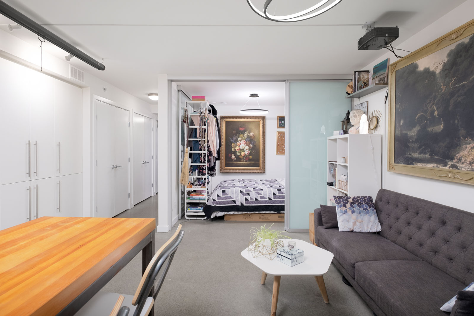 Modern studio apartment with a visible bedroom, kitchen, and living area; artwork and shelves decorate the walls, and a wooden dining table is in the foreground.