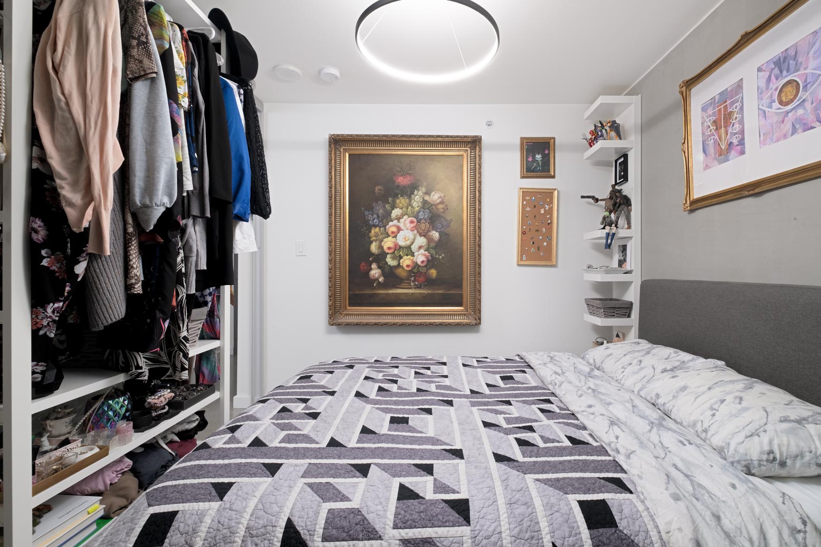 A neatly organized small bedroom with an open wardrobe, geometric patterned bedding, a large floral painting, and framed art on the walls.