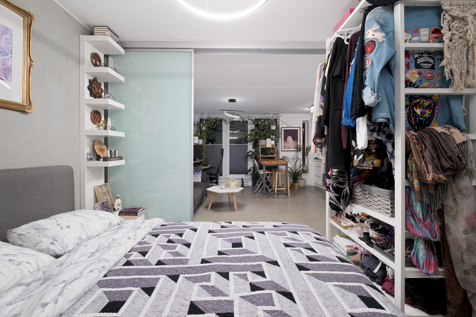 A modern bedroom with a patterned bedspread, open closet, and shelves, looking into a living area with plants, a table, and chairs through sliding glass doors.