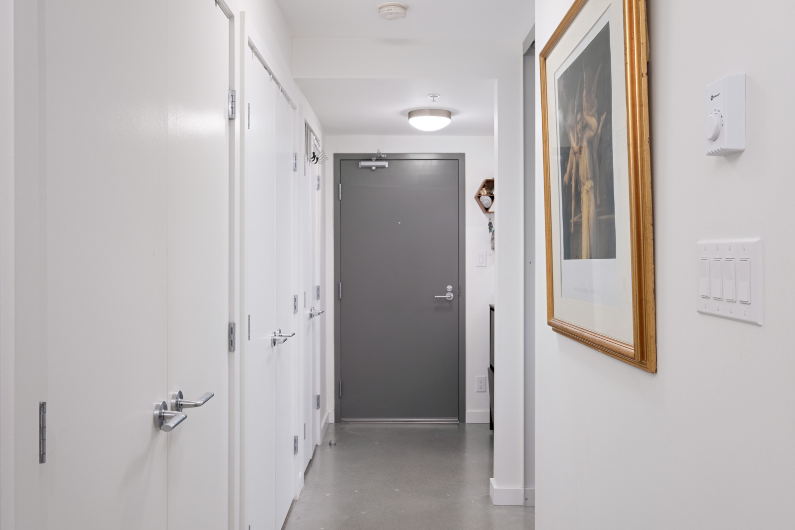 A clean, minimalist hallway with white walls and doors, a gray entry door at the end, a ceiling light, and a framed artwork on the right wall.