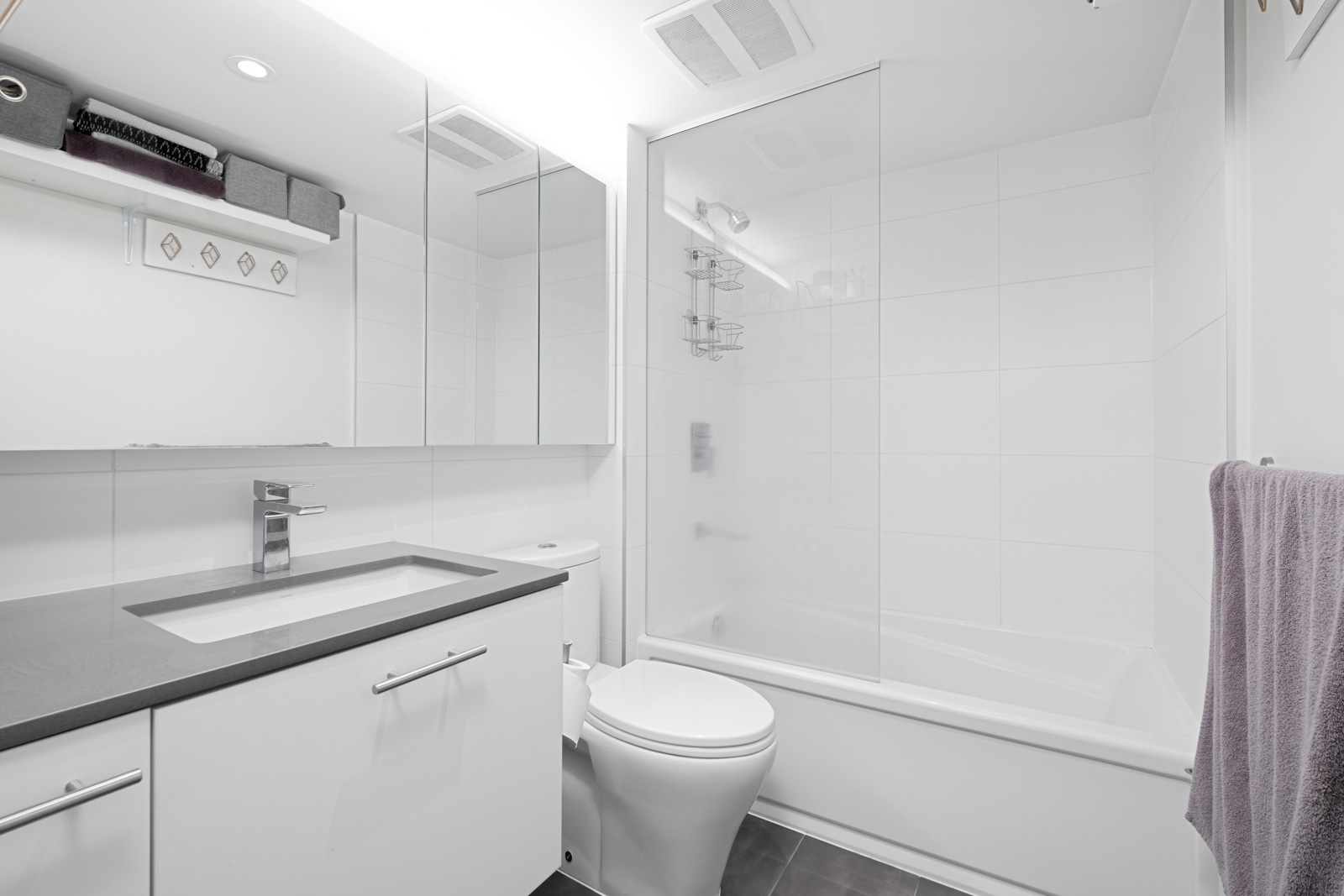 Modern white bathroom with a large mirror, gray countertop, sink, toilet, towel rack, shelves, and a bathtub with a glass shower screen.