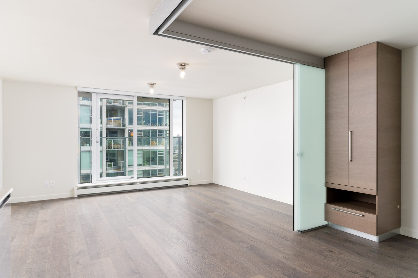 Empty modern apartment interior with a large window, sliding glass partition, wooden floor, and built-in cabinet.