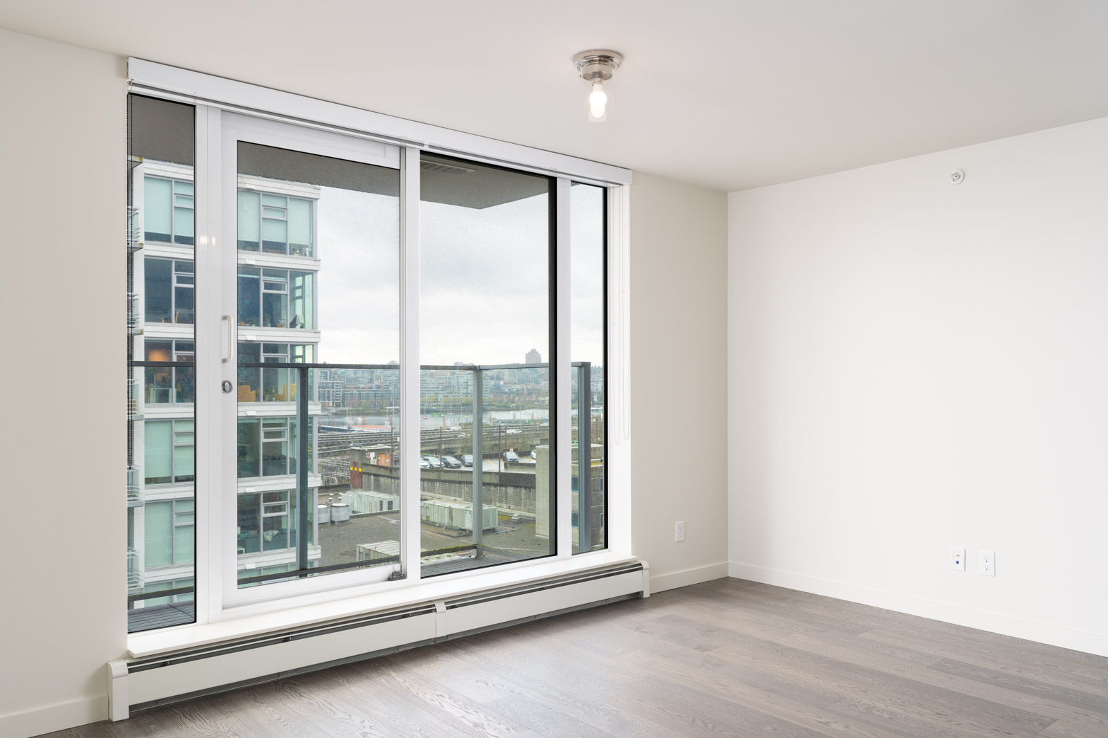 Empty room with light-colored walls, hardwood floor, and a large sliding glass door leading to a balcony. View of modern buildings and a cityscape in the background.