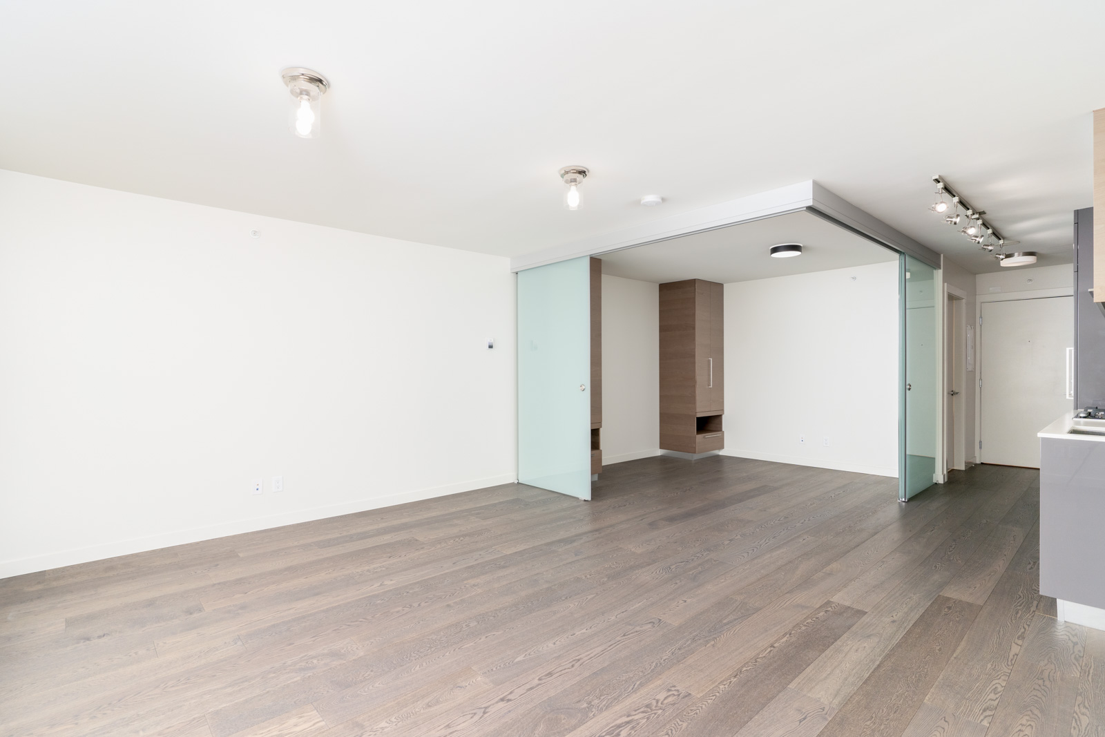 An empty modern room with hardwood floors, sliding glass doors, recessed lighting, and a small kitchen area on the right.