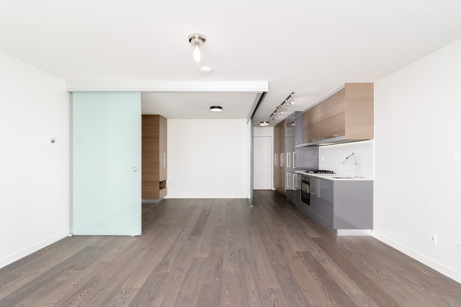Bright, empty modern kitchen with wooden floors, sleek cabinets, built-in appliances, and a sliding glass door. Minimalist design with white walls and recessed lighting.