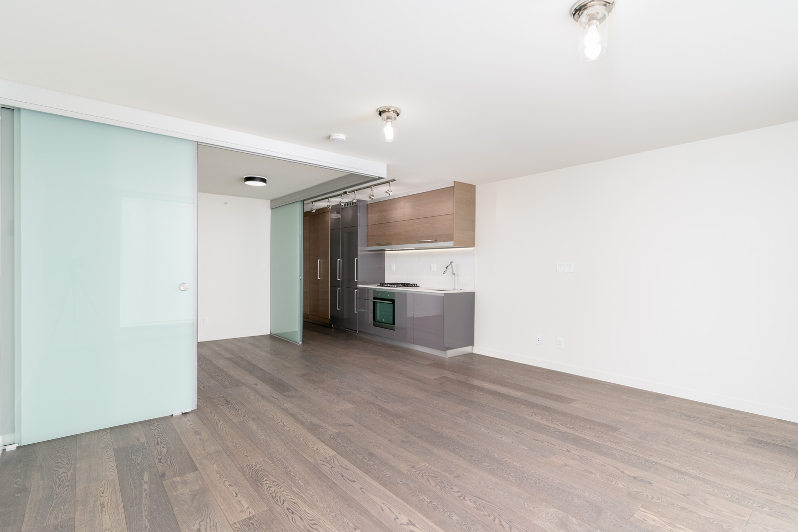 A modern, empty apartment interior with wooden floors, a small kitchen area with appliances, and frosted glass sliding doors.