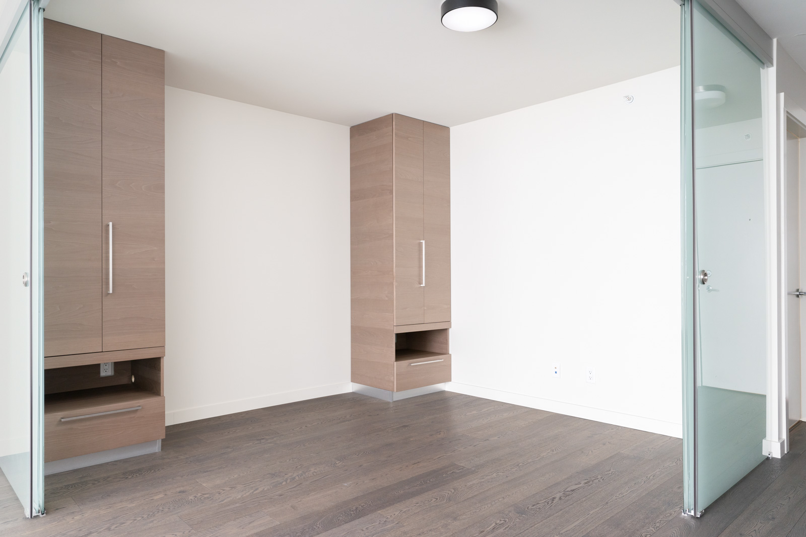 Minimalist room with light wood cabinets, gray hardwood floor, and glass partition walls. Ceiling light fixture in the center.