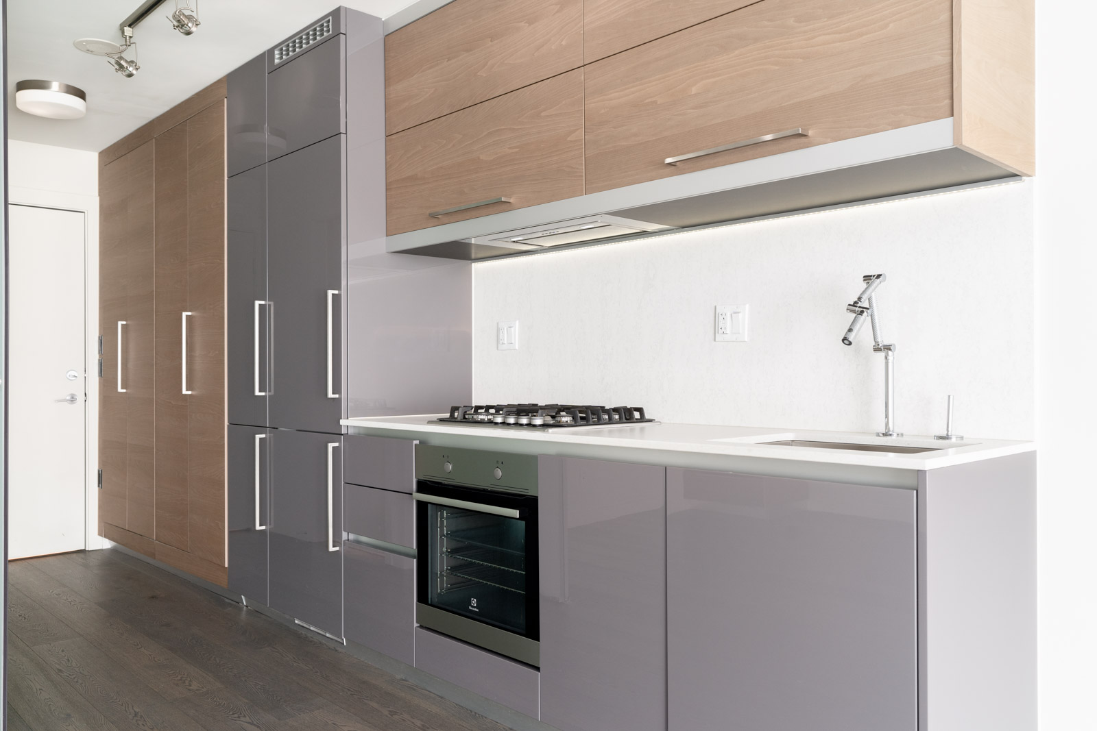 Modern kitchen with built-in gray appliances, wood cabinets, and under-cabinet lighting. Includes a stovetop, oven, and sink with a sleek faucet on a minimal white countertop.