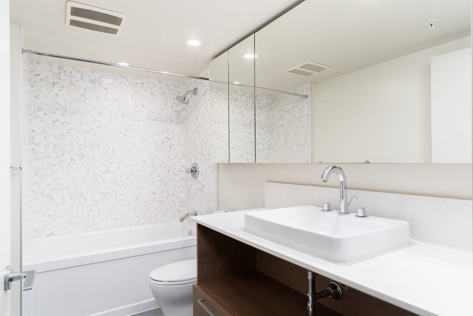 Modern bathroom with a rectangular sink, white countertop, and wooden cabinet. A bathtub with a tiled backsplash and a large mirror above the sink are visible.