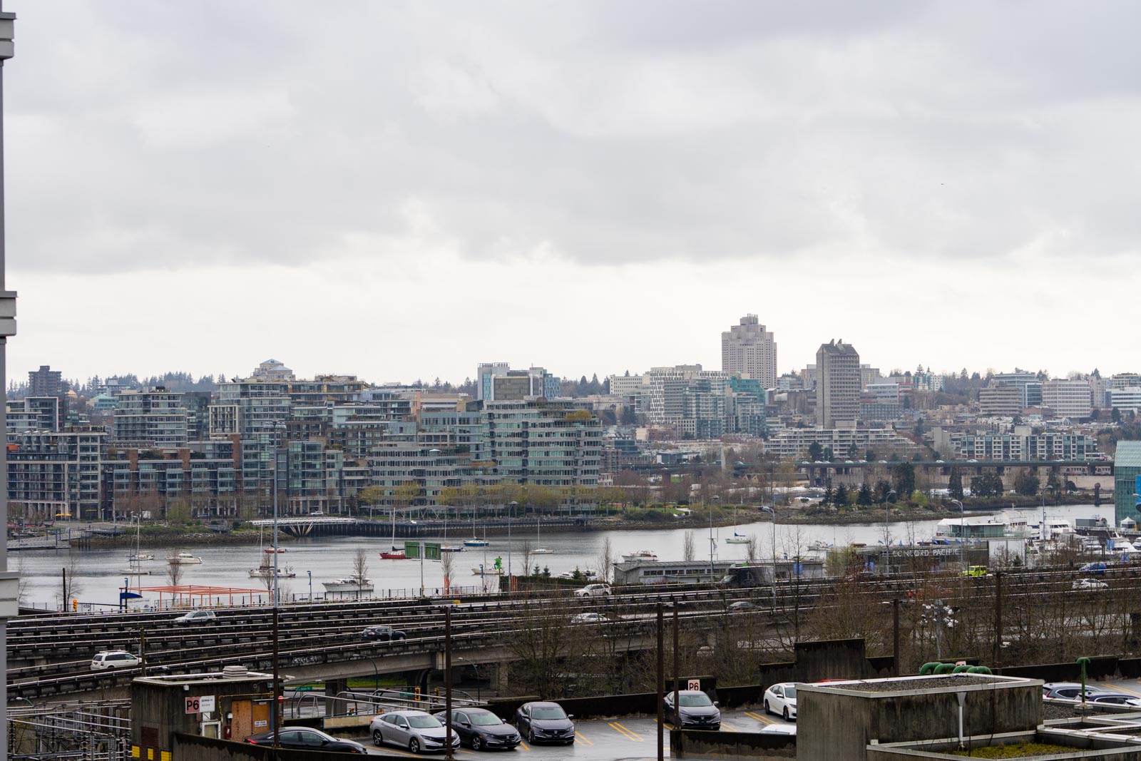 Urban skyline with waterfront, low-rise buildings, and a cloudy sky. Cars are parked in the foreground, and boats are on the water.