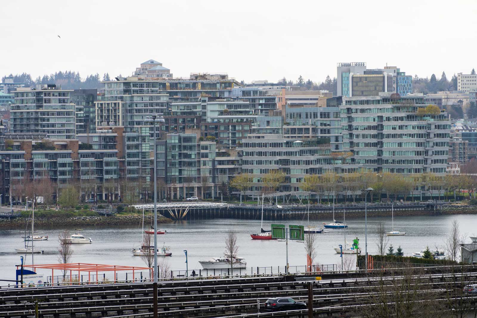 Cityscape view of a waterfront with modern high-rise buildings, boats on the water, and a rail line in the foreground.