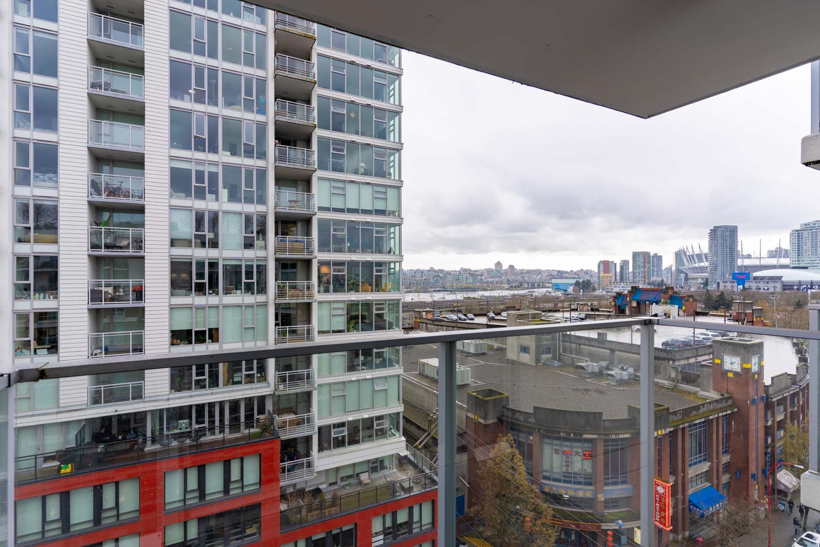 View of a cityscape featuring a modern apartment building with glass balconies in the foreground and a cloudy sky above.