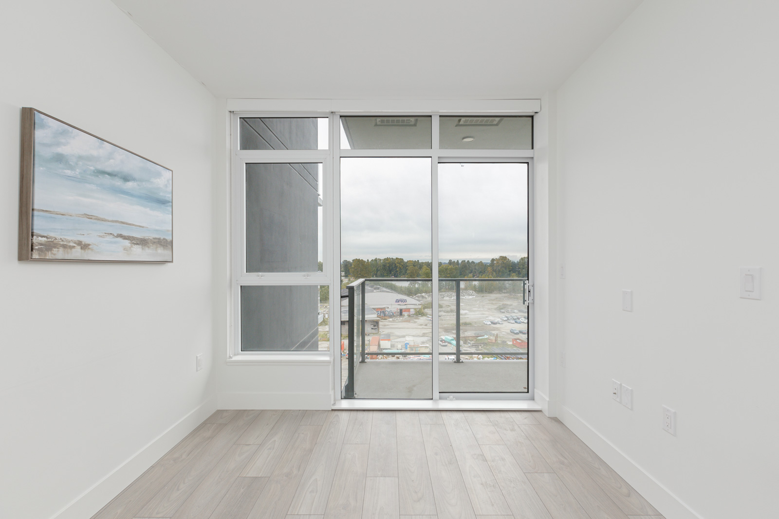 Empty room with light wood flooring, a large window, and a glass door leading to a balcony. A landscape painting is on the wall. The view shows a cloudy sky and construction site.