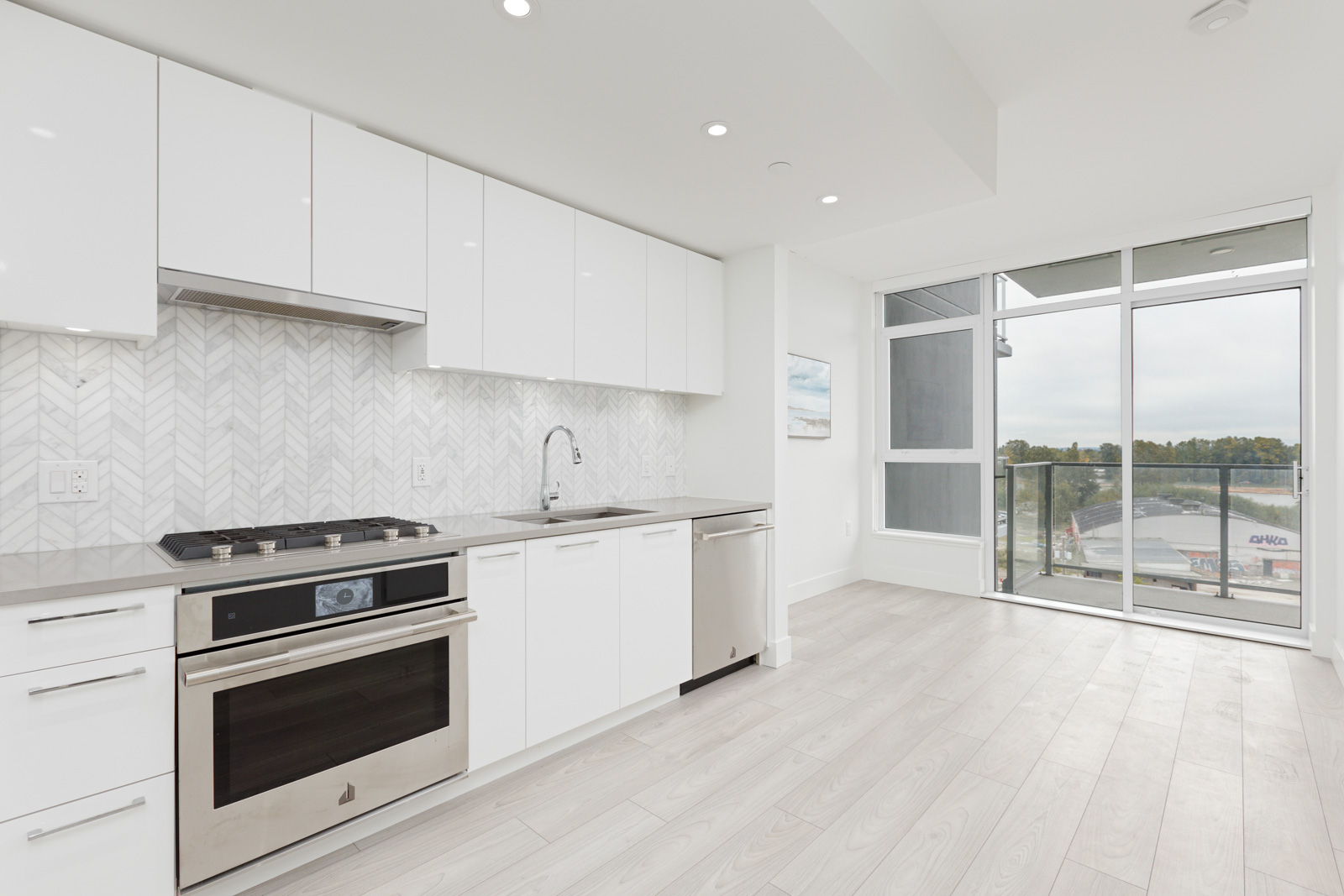 Modern kitchen with white cabinets, stainless steel appliances, and herringbone backsplash. Large window with a view of trees and buildings. Light wooden floor.