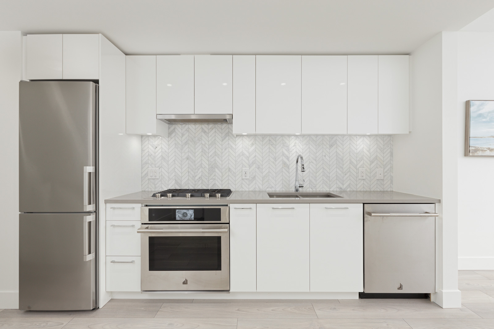 Modern kitchen with white cabinets, stainless steel refrigerator, oven, and dishwasher. Herringbone tile backsplash and light wood flooring.