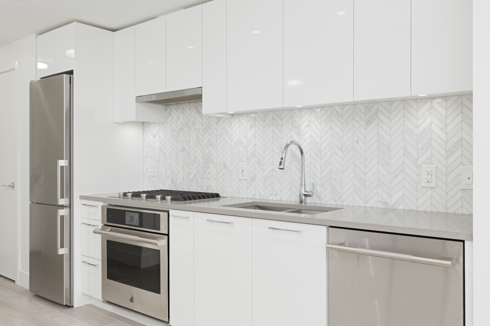 Modern kitchen with white cabinets, stainless steel refrigerator, oven, dishwasher, and a sink beneath a geometric-patterned backsplash.