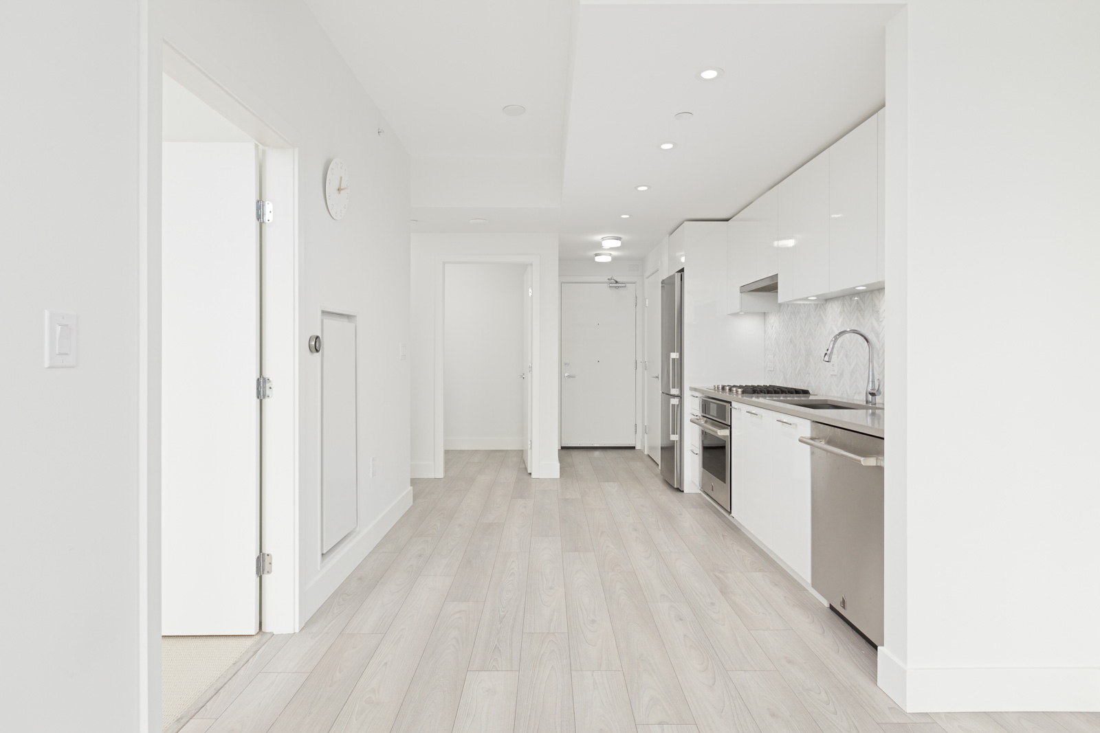 Minimalist kitchen space with white cabinets, light wood flooring, a built-in oven, and stainless steel appliances. There is a clock on the wall and a hallway leading to a door.