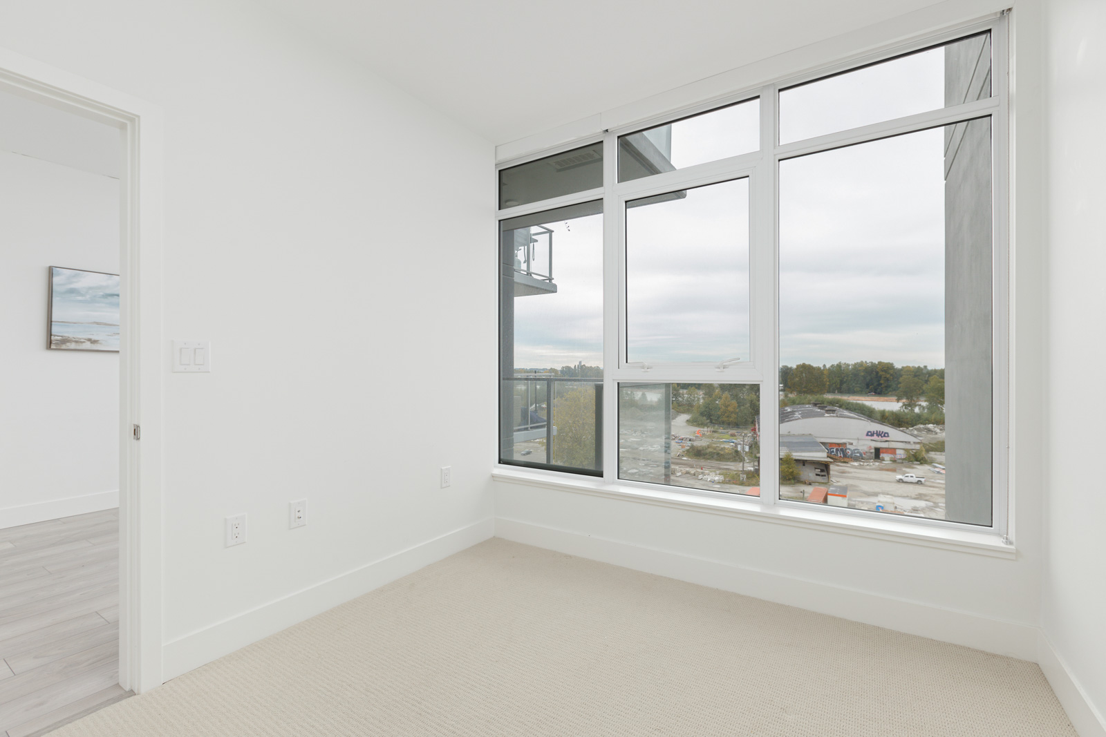 Empty room with large windows, beige carpet, and white walls, offering a view of a construction site and trees outside. Adjacent room visible.