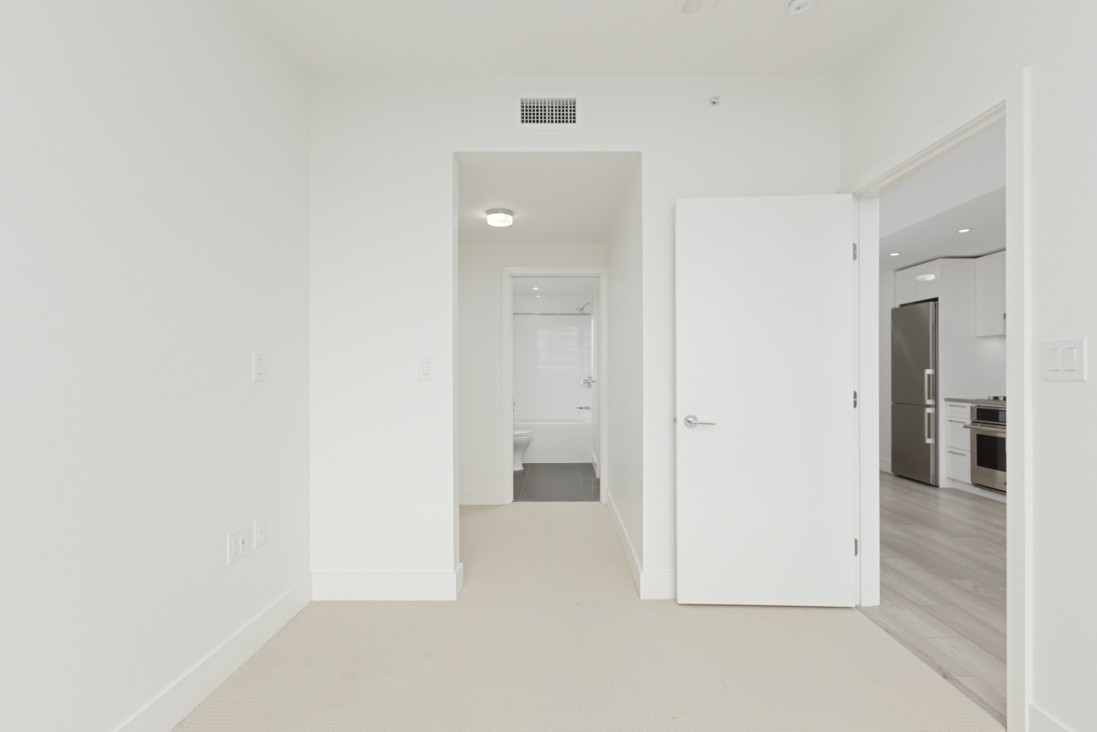 Empty room with beige carpet, open door leading to a kitchen area, and another door to a bathroom. Walls and doors are white. Ceiling light is visible.