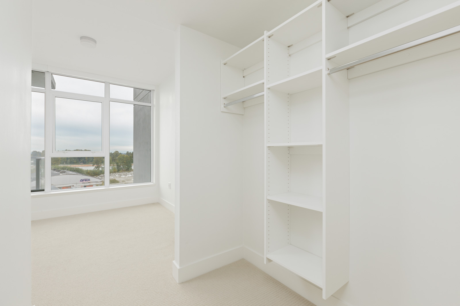 Spacious, empty walk-in closet with white shelves and clothes rods. Large window with a view outside, adjacent to beige carpeted floor.