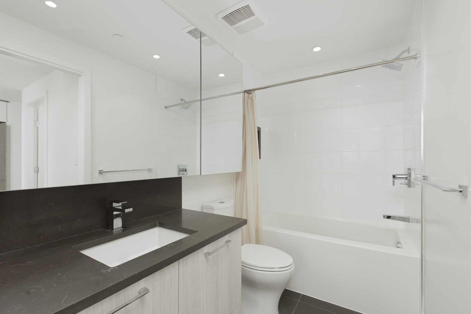 Modern bathroom with black countertop, sink, and mirror above. White bathtub with curtain, toilet, and recessed lighting complete the minimalist design.