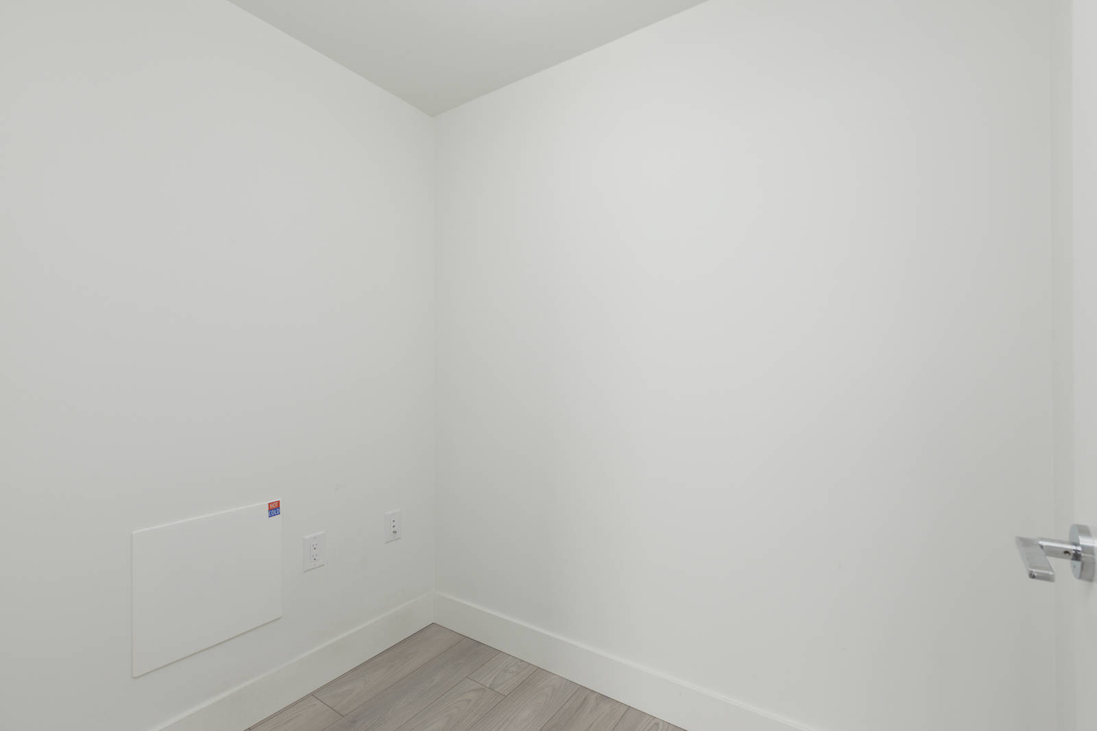 Empty white room with a small electrical panel and outlet on the wall. Light wood flooring and closed door visible.